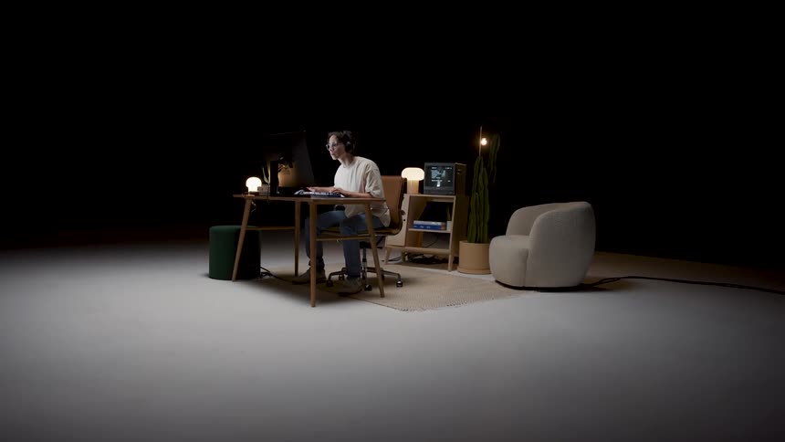 A man wearing headphones is sitting at a desk typing on a computer keyboard and there is living room furniture in a wide open room