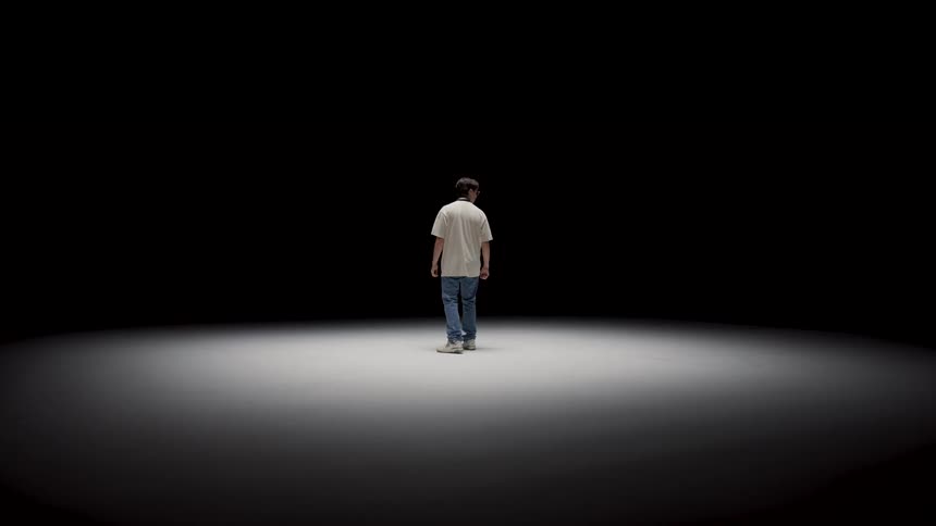 A man stands in an empty room dark room with a spotlight and looks up amd around