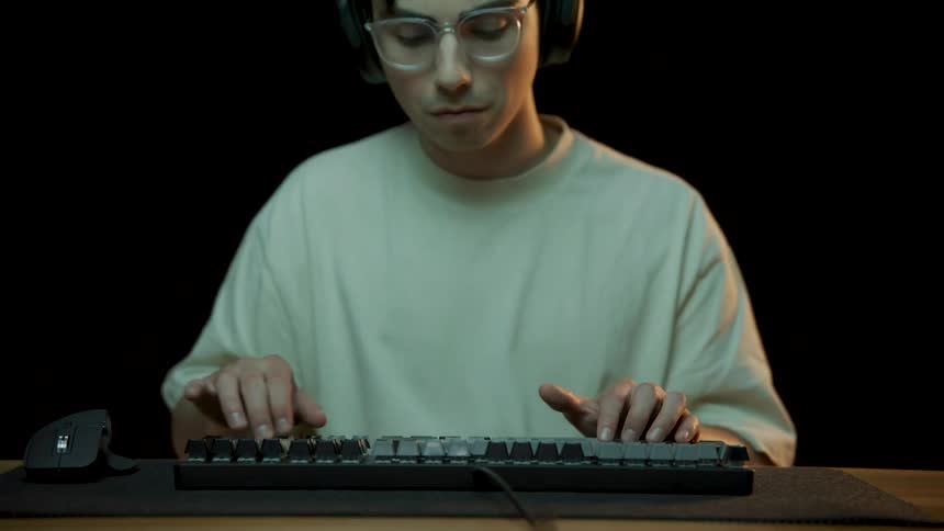 Man wearing glasses headphones and white t shirt taps on keyboard sitting at desk in room