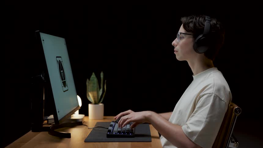 Man wearing glasses headphones and white t shirt sits at desk working on computer in dark room
