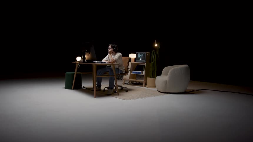 Man with headphones glasses and white t shirt sits at desk watching computer screen in dark room