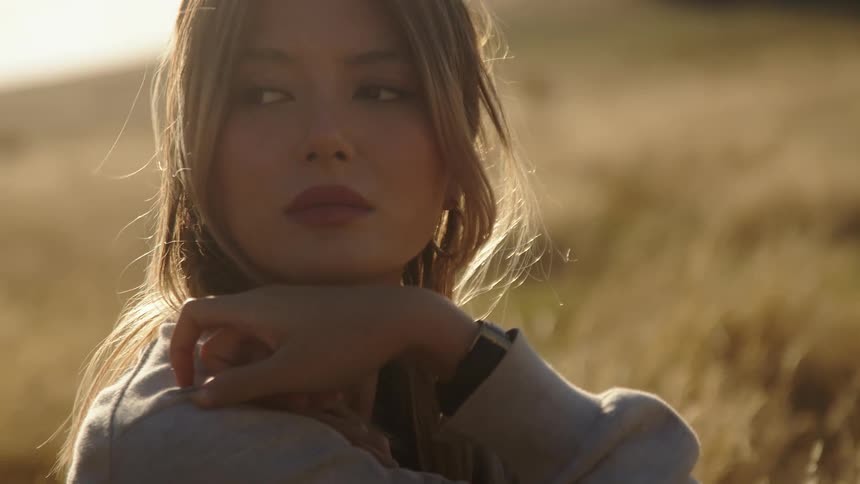 A young woman is sitting and relaxing in a sunny field with her hand on her shoulder