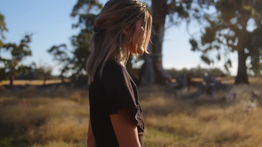 Woman with fair hair a pair of white shoes black skirt and shirt walks across the field near a clump of trees