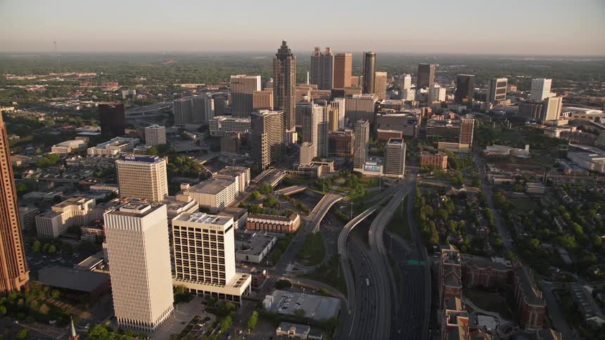 A aerial video captures the view of Downtown Connector towards downtown Atlanta revealing immense skyscrapers and towers overlooking the vibrant and busy streets during a hazy and sunlit day