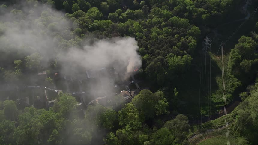 Approaching aerial footage of smoke rising from a residential house fire in a wooded area during daytime in West Atlanta Georgia
