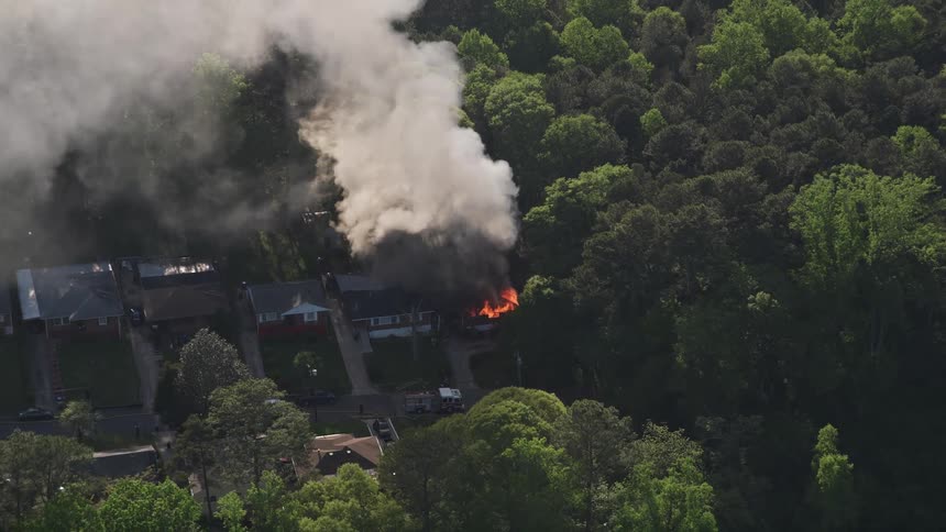 Aerial footage approaches and tilts down to reveal a birds eye view of a house engulfed in flames and smoke in West Atlanta Georgia with fire trucks on the street below