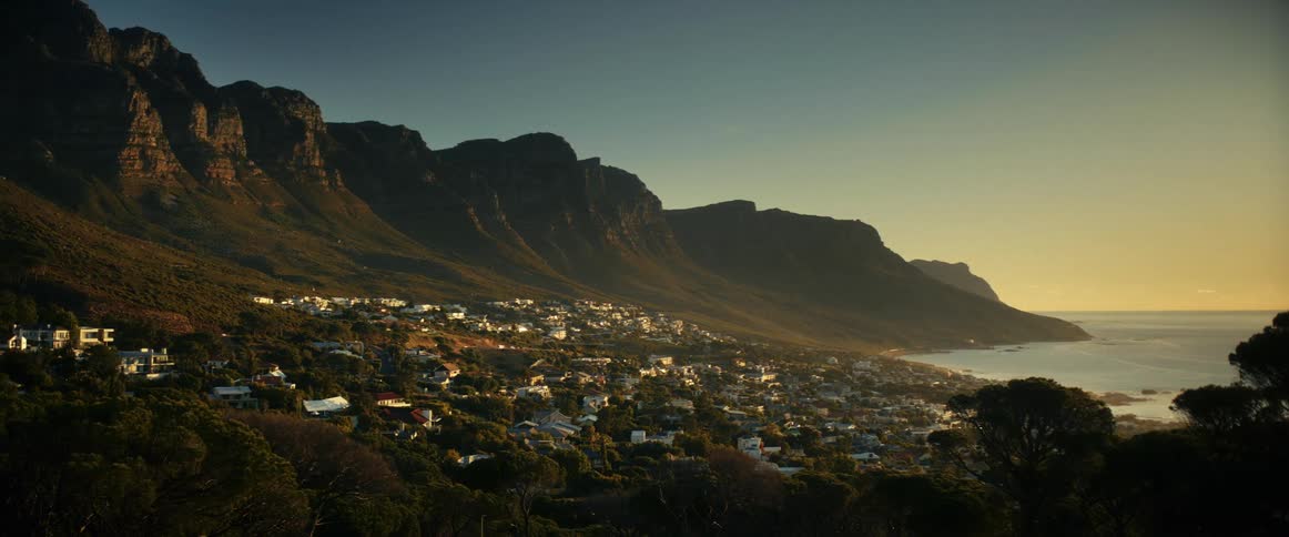 Traffic moves on a road through a mountainside community along the coast as waves roll in over the beach as the sun sets in the distance