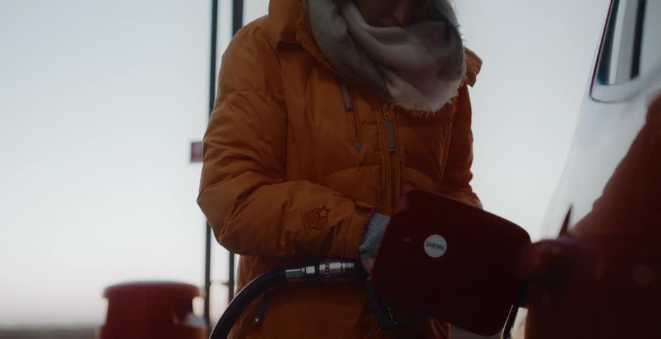 Woman with orange coat brown scarf and gray hat fills up gas tank of car at gas station