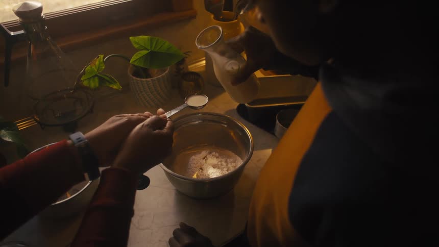 A man and a woman are in the kitchen standing over a silver bowl of ingredients as they measure a spoonful of milk and pour it into the bowl