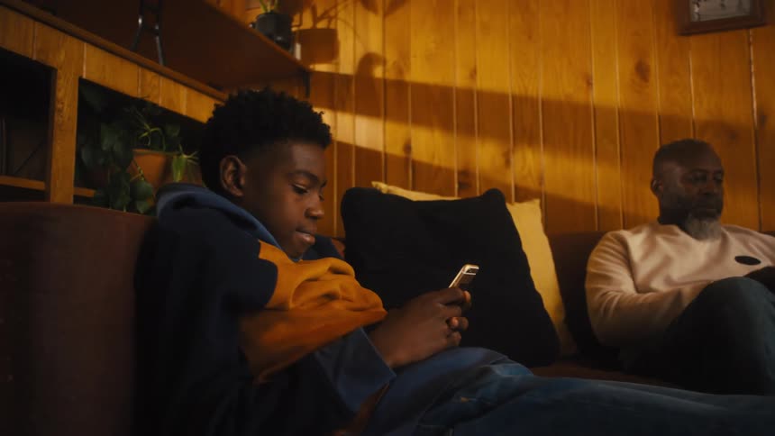 A man and boy uses a phone and tablet on a living room sofa