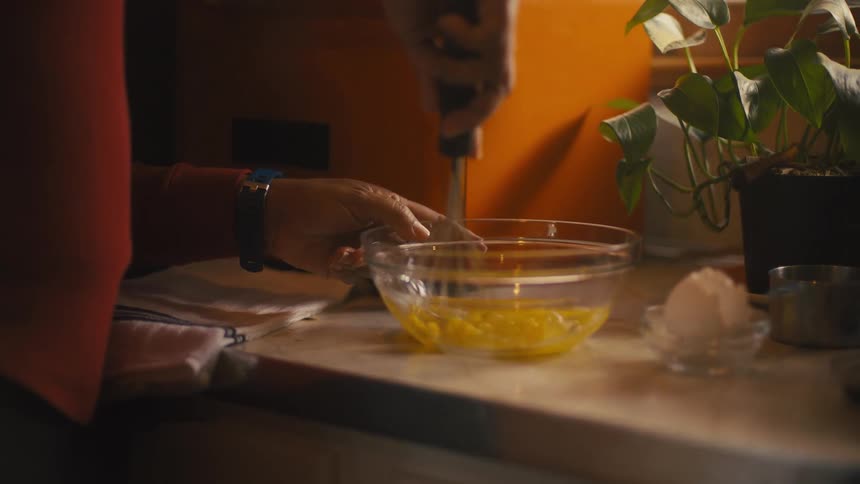 A woman is standing at the kitchen counter beating eggs in a clear bowl with a wire whisk