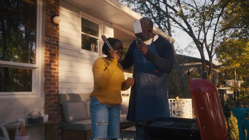 A man and his daughter hug and clink cooking utensils together as they stand in front of a grill