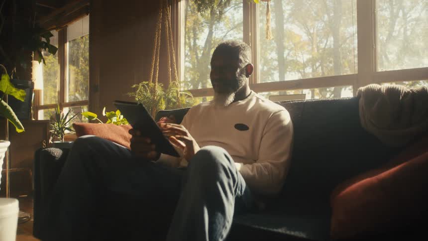 Older man sitting on couch near wall of windows smiles as he views tablet