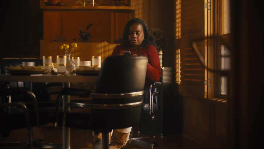 A black woman sit at a table in a room and looks down at her phone