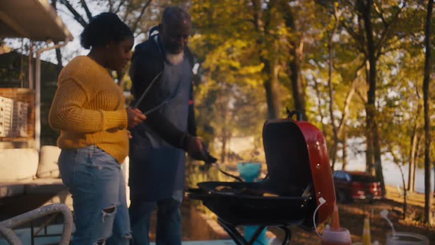 A father and teenage daughter cooking hot dogs and hamburgers on an outdoor grill in autumn