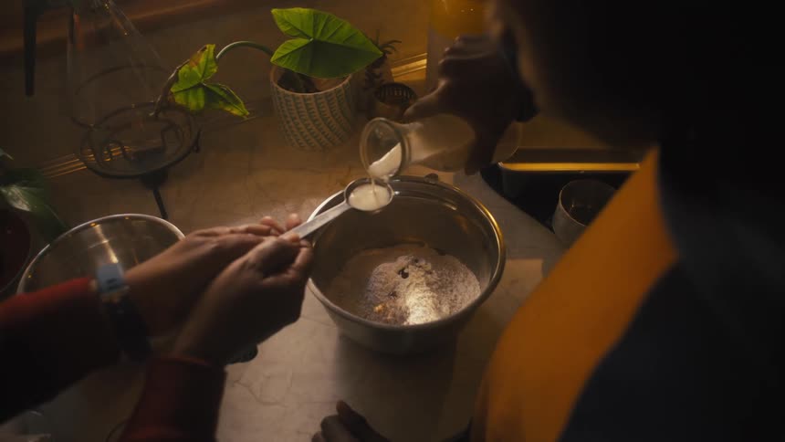 A man and a woman are in the kitchen standing over a silver bowl of ingredients as they measure a spoonful of milk and he pours it into the bowl