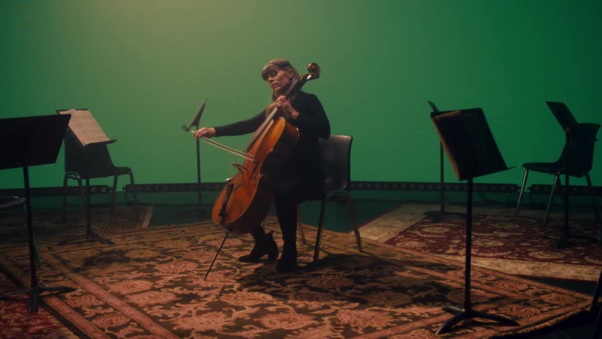 A woman plays the cello while seated in front of a green screen