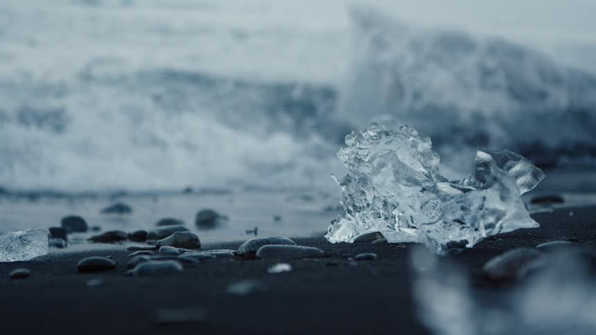 Surf flows over large block of ice on edge of beach before continuing to spot small ice chunk lies