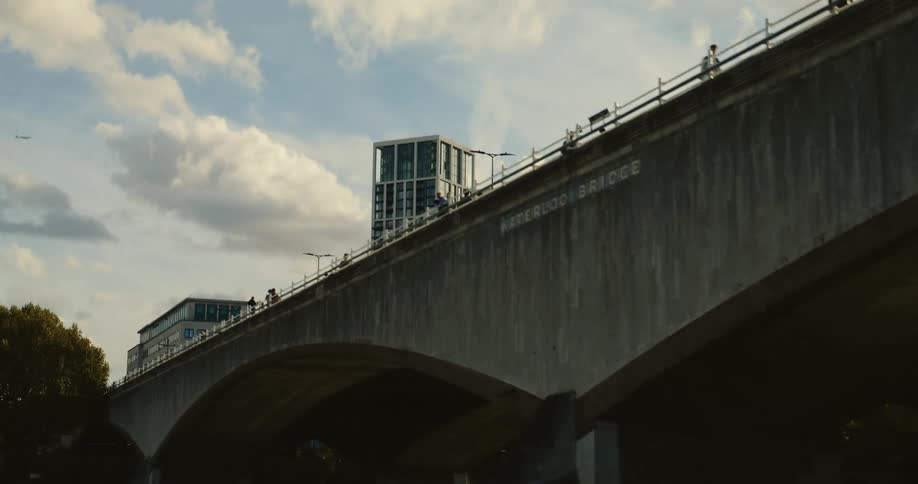 People are walking across Waterloo bridge near the city with tall buildings in the distance under a cloudy sky
