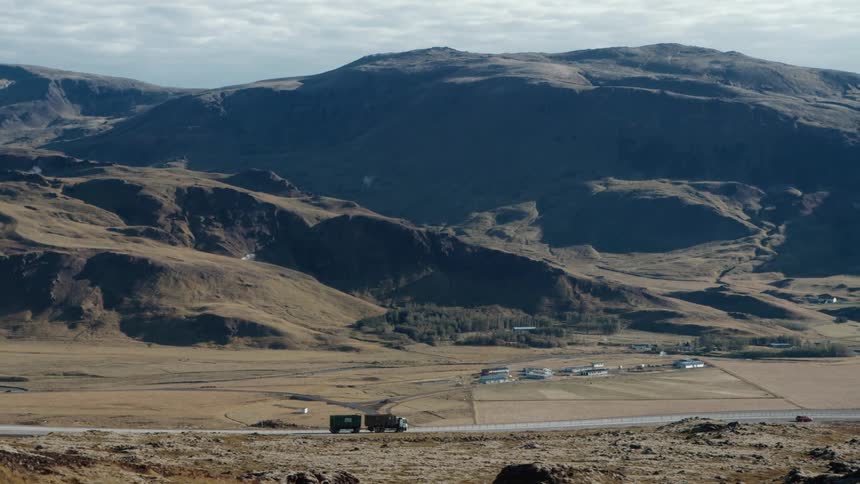 A truck and a car drive on a road near a mountain range and pass by a cluster of buildings