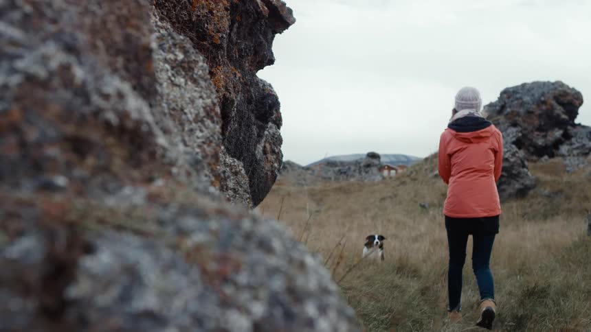 A woman in a coat and knitted cap walking with her pet dog near rock structures