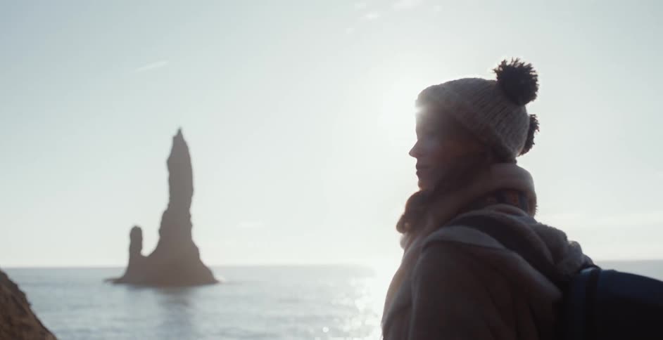 A woman stands along the coast and looks at large rocks
