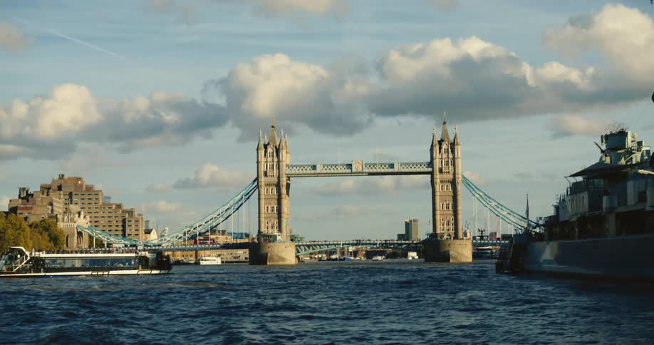 Several people ride ferry boat along river near tower bridge under a cloudy sky
