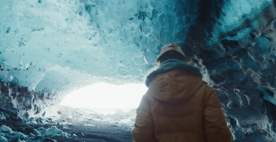 A person wearing a yellow winter coat and a hardhat is walking out of a frozen cave as water drips from melting ice