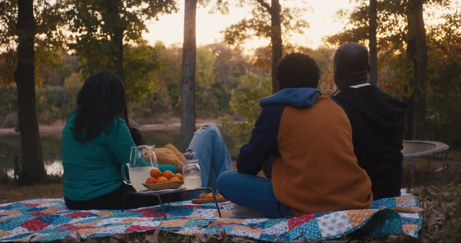 A family is sitting on a picnic blanket with snacks looking at a lake