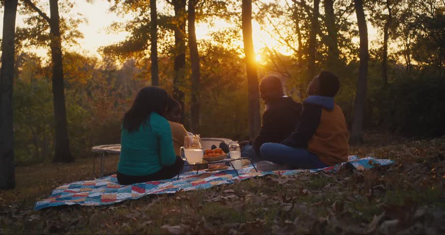 A family is sitting on a blanket outside in their yard near a trampoline having a picnic and watching the sun set through the trees