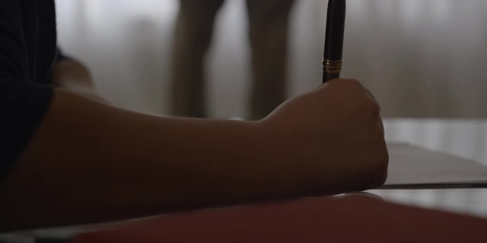 A young child sits at a desk in school and quickly writes on a paper as a teacher stands back and watches