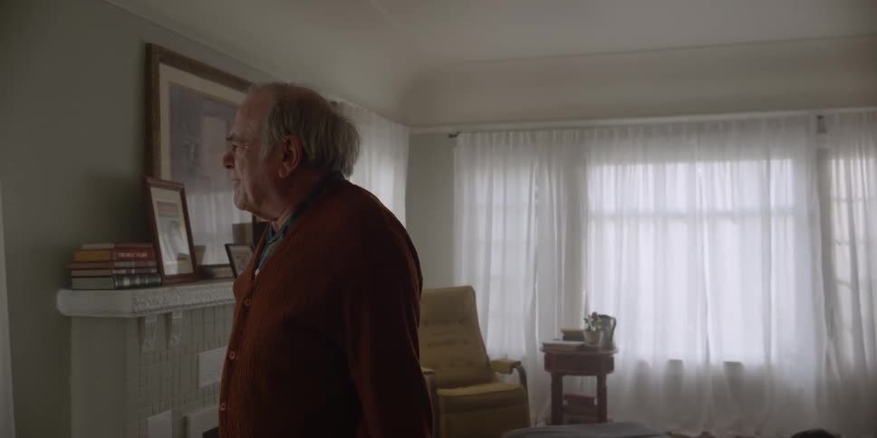 A man walks across his living room as he speaks and moves his hands as he turns around and goes back