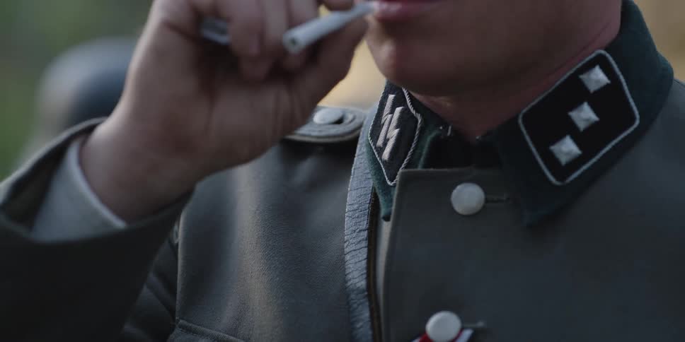 A man in a military uniform puts a cigarette in his mouth and lights it with the lighter in his hand
