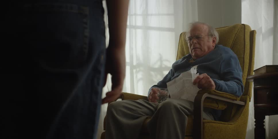 A man is sitting in a chair in his living room holding papers in one hand and a glass of water in the other hand as he speaks to someone and they walk toward him