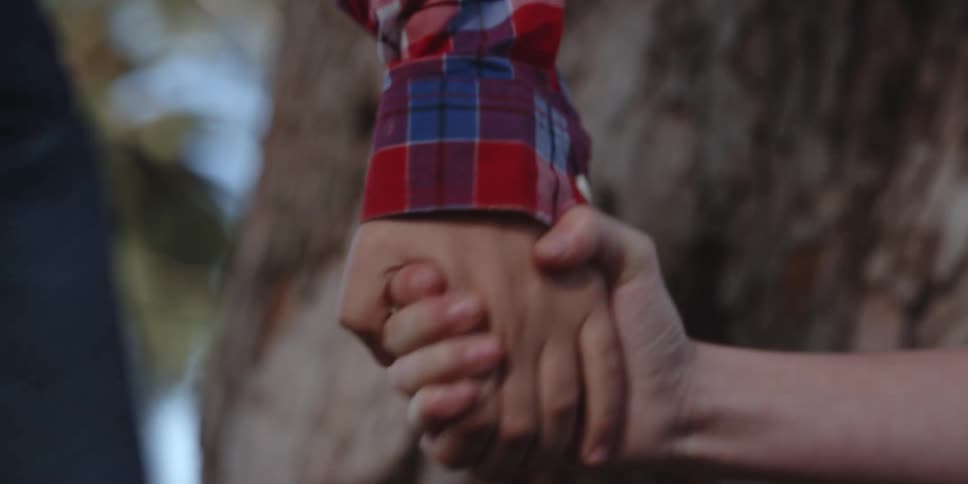A boy walks to a tree and puts his hand out and a girl raises hers