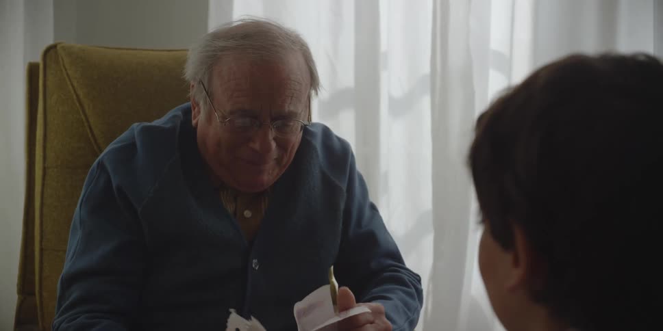Grandson handing grandfather wearing glasses and dark blue sweater a piece of paper sitting in armchair
