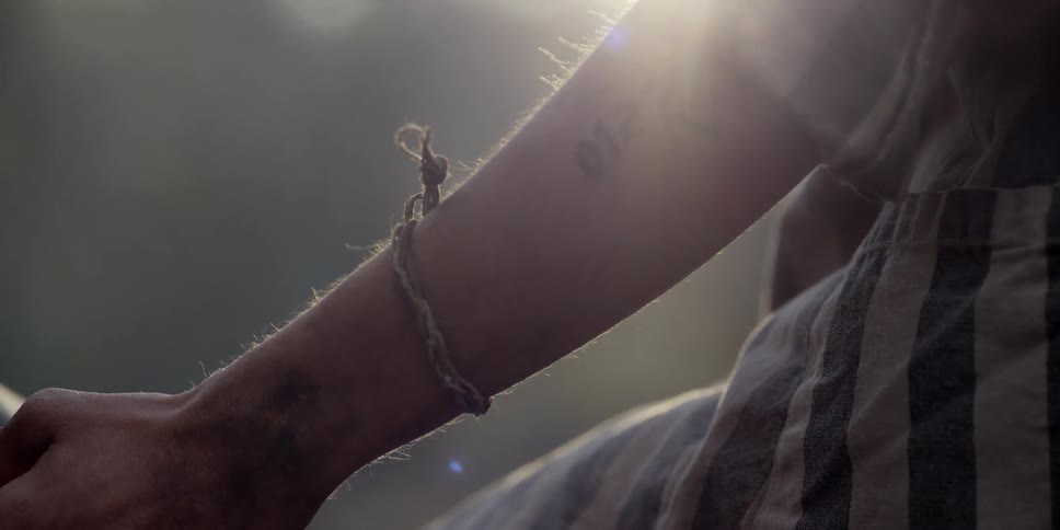 A man ties a bracelet on another mans arm as the sun shines brightly in the distance