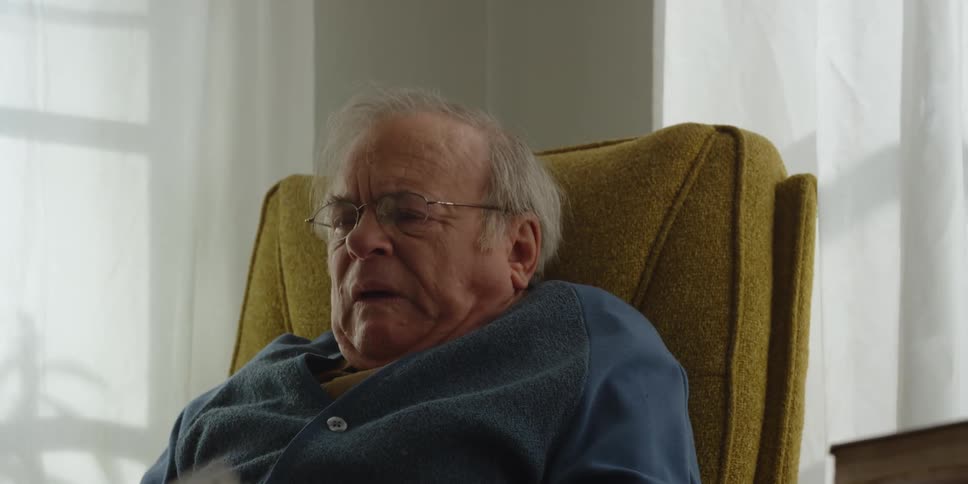A man sits down in a chair with a glass of water and a sheet of paper with writing on it as he takes off his glasses and looks up at the person in front of him