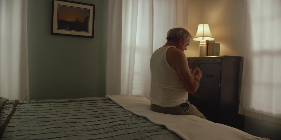 A man is sitting on the edge of his bed looking at papers and he takes off his glasses and looks across the room