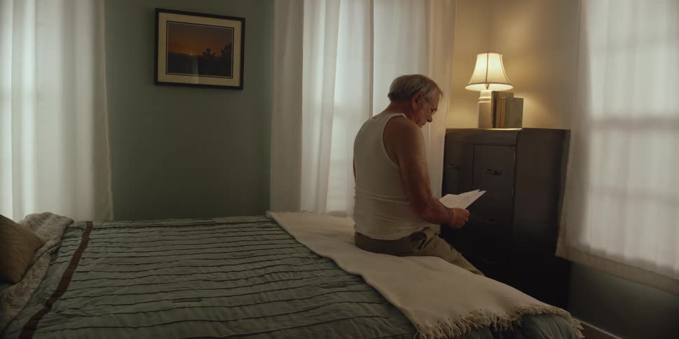 Elderly man wearing glasses white under shirt and brown pants sits on bed in bedroom