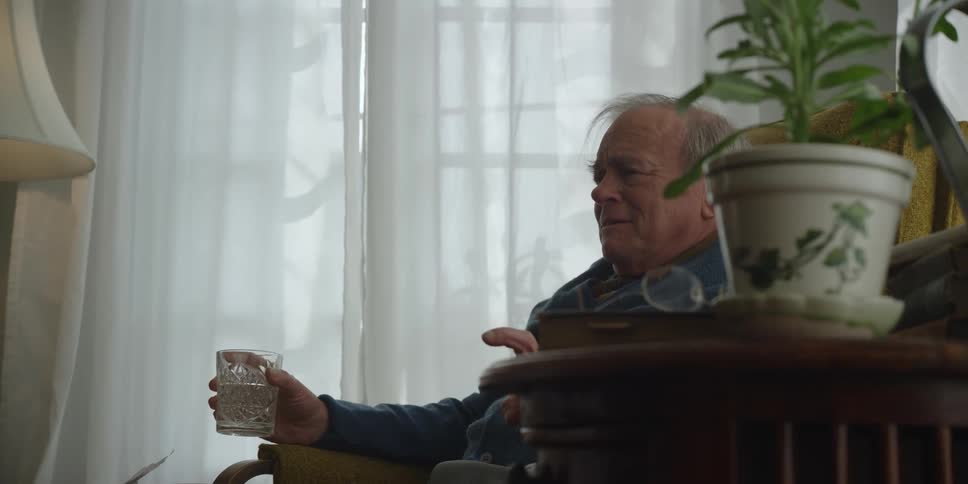 A man is sitting in a chair in his living room holding a glass of water and he sets it down on the table next to him