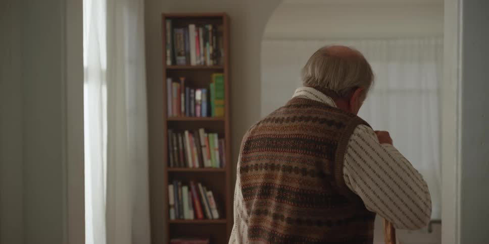 A man is standing in his living room holding a cup of coffee and he turns and takes a drink as he walks away