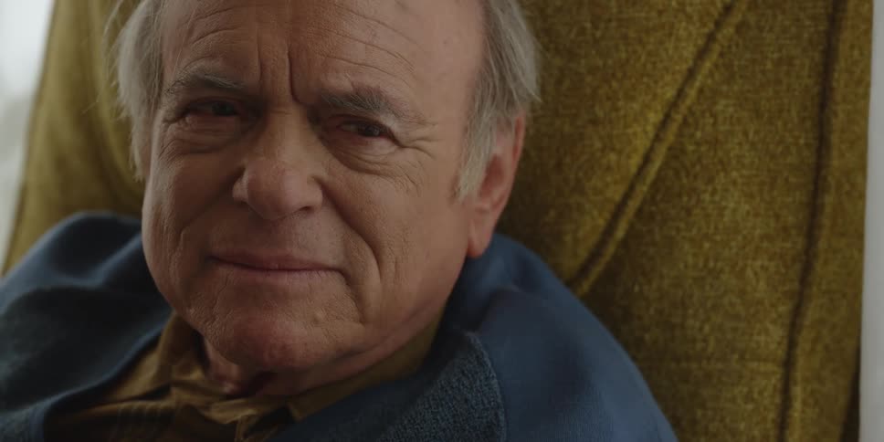 An elderly man sits in a chair and smiles
