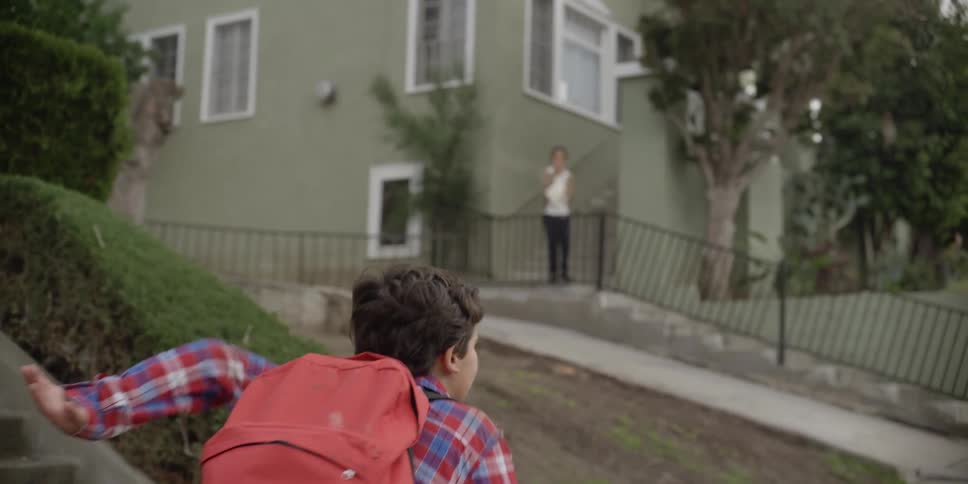 A boy standing on a sidewalk wearing a backpack starts to run toward a building