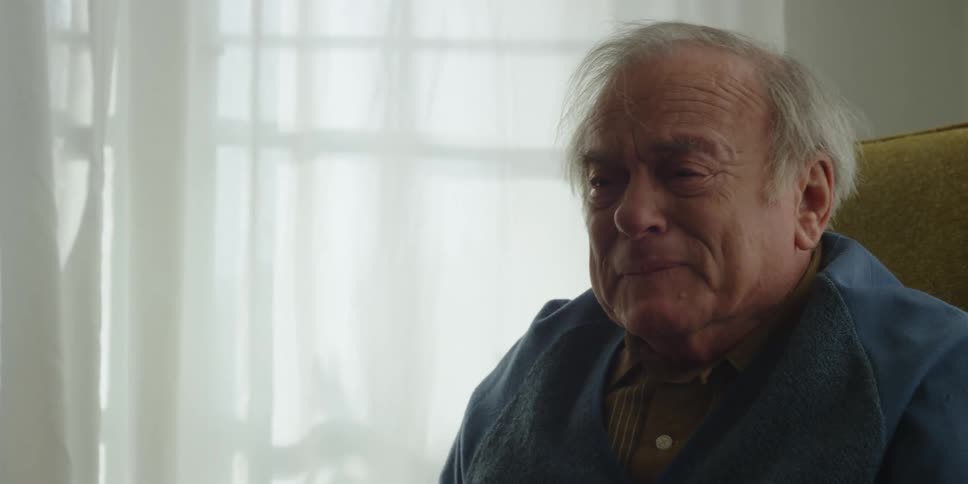 An elderly man smiling and getting sad while sitting in a living room chair