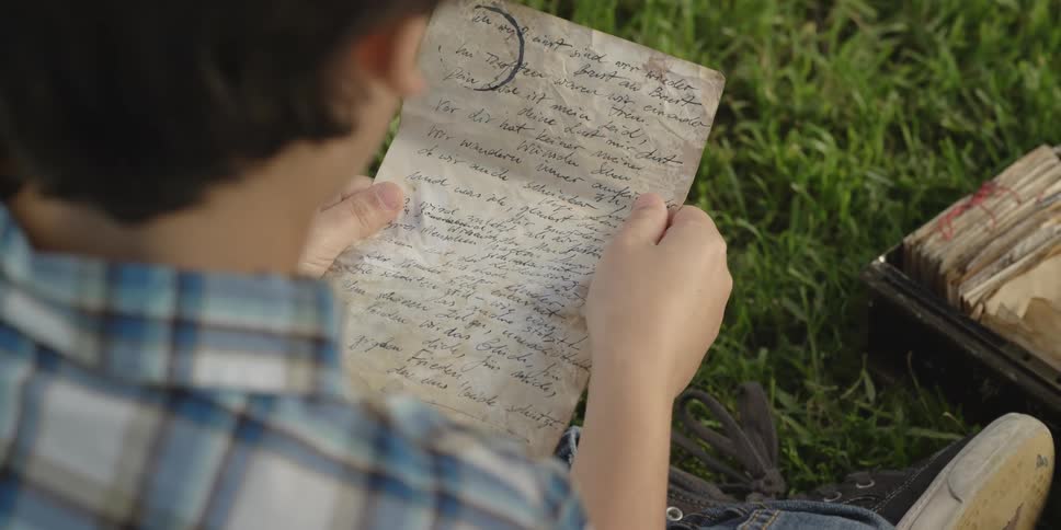 A young boy sits in the grass and reads an old hand written note next to a woman holding a cell phone who looks through a box of letters