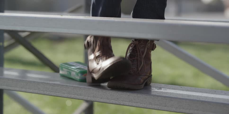 A person in brown lace up boots sitting outside on metal bleachers