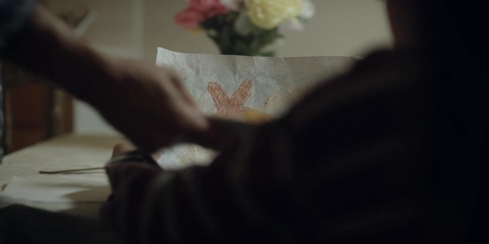 A boy is sitting at the table holding a drawing when his mother puts food on the table and sits down
