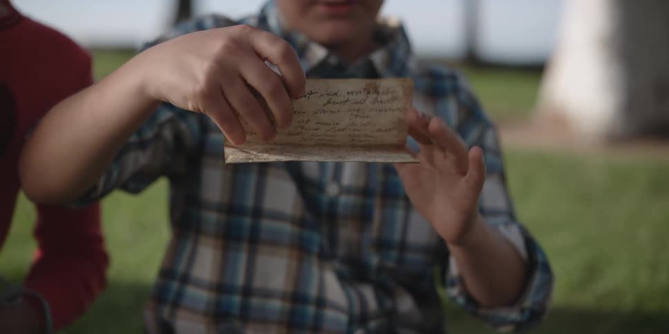 A young boy sits on the grass outside with a girl and picks up an old hand written note and unfolds it