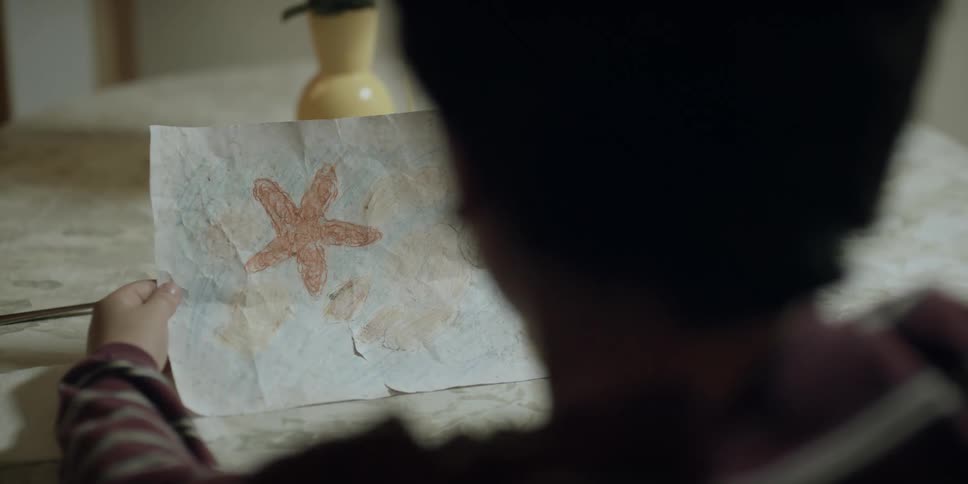 A boy is sitting at the table holding a drawing when his mother puts food on the table and sits down